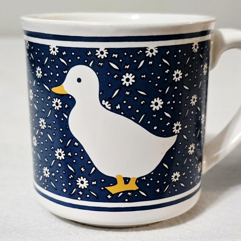 May include: A white ceramic mug featuring a dark blue background decorated with a repeating pattern of small white flowers. A large, solid white duck with a yellow beak and yellow feet is centered on the front of the mug, which has a handle.