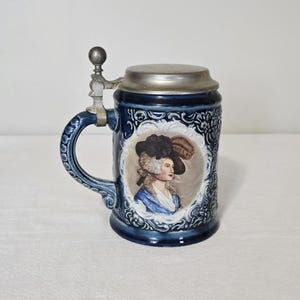 May include: A dark blue ceramic beer stein with a silver lid and handle. The stein features a portrait of a woman in a hat and a decorative floral pattern. The handle is ornate, and the lid is hinged.