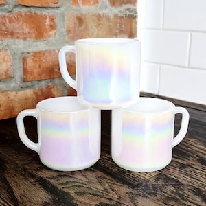 May include: Three white glass mugs with a smooth, iridescent finish that reflects soft rainbow colors. Each mug features a rounded body, a curved handle on the side, and a glossy surface that catches the light, creating a pearlescent effect.