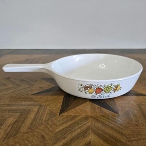 May include: White ceramic skillet with a white handle. The skillet has a floral design with a red pepper, green pepper, and other vegetables. The text "Le Persil" is printed on the bottom of the skillet.