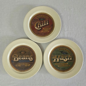 May include: Three off-white plates with vintage-style labels. One plate reads "Aunt Jenny's Chili", another "Aunt Jenny's Beans", and the third "Aunt Jenny's Hash". Each label has a brown, red, and green color scheme with text that reads "Since 1895".