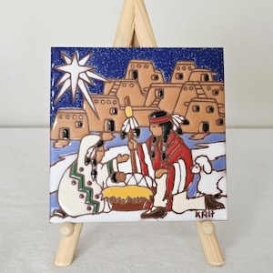 May include: Square ceramic tile featuring a nativity scene with Native American figures. The tile has a blue sky background with a star, adobe-style buildings, and figures in traditional clothing. The tile is displayed on a small wooden easel.