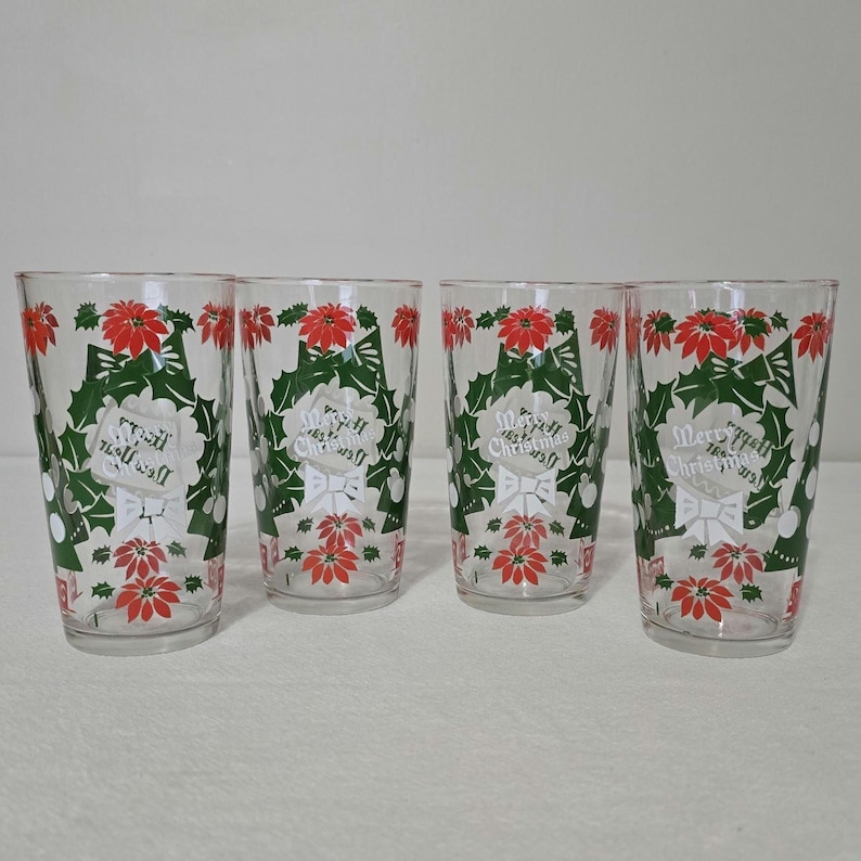 May include: Four clear drinking glasses with a Christmas theme. Each glass features a design of red poinsettias, green holly leaves, and the words "Merry Christmas" in a white banner. The glasses are cylindrical and appear to be made of glass.