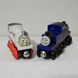 May include: Two wooden toy trains. One is white with red wheels and a smiling face. The other is blue with a black top and a smiling face. Both trains have black wheels and are on a white surface.