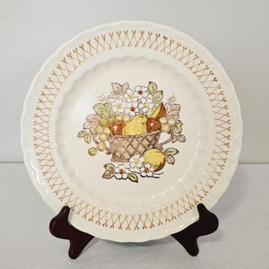 May include: Off-white ceramic plate with a scalloped edge and a brown crosshatch pattern. The center features a basket of fruit in shades of yellow, green, and brown, surrounded by white flowers and green leaves. A decorative piece for the kitchen.