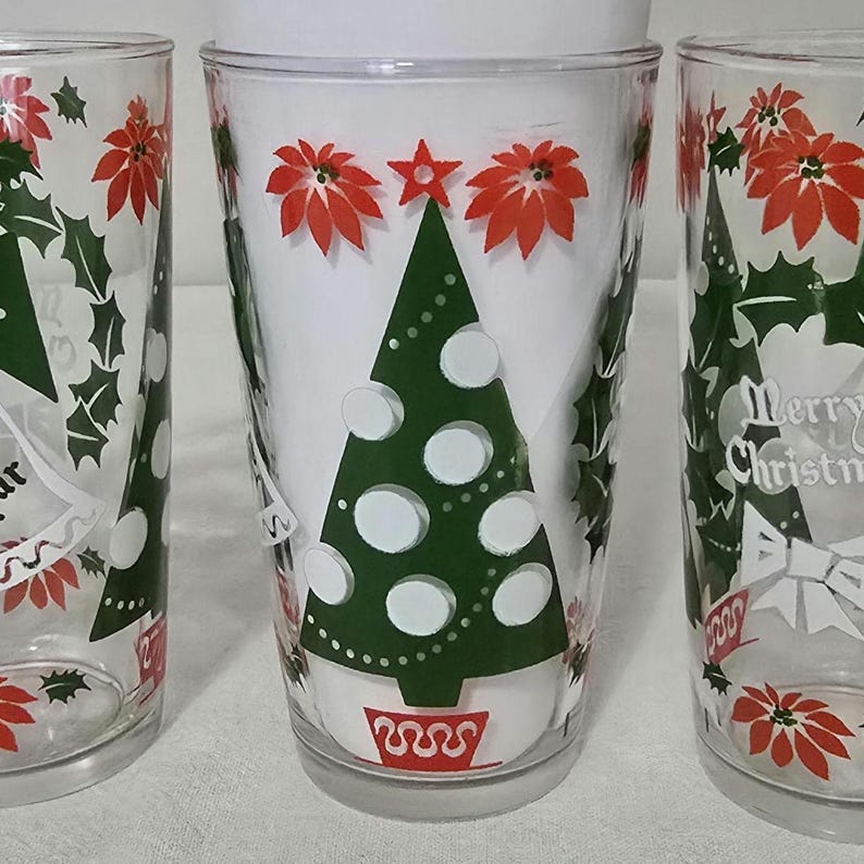 May include: Three clear drinking glasses with Christmas-themed designs. Each glass features a green Christmas tree, red poinsettias, and the words "Merry Christmas" in white. The glasses are decorated with holly and star accents.