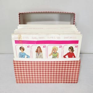 May include: A red and white gingham storage box containing a set of sewing pattern index cards. The cards feature illustrations of various clothing styles labeled Classic Styles, Flared Skirts, Empire Styles, and Shirtdresses with codes A1 to A4.