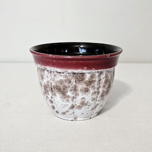 Peut inclure: Un cache-pot en céramique avec un bord bordeaux profond et un corps blanc orné d'un glaçage moucheté brun et gris. L'extérieur présente une texture subtilement côtelée verticalement, tandis que l'intérieur est fini en brun foncé brillant.