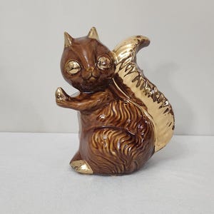 May include: A brown ceramic squirrel figurine with a gold-painted tail. The squirrel has large, round eyes and a playful expression.