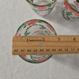 May include: Clear glass Christmas-themed shot glasses with red poinsettias and green holly leaves. The glass is approximately 2 inches in diameter. The glasses are made in China.