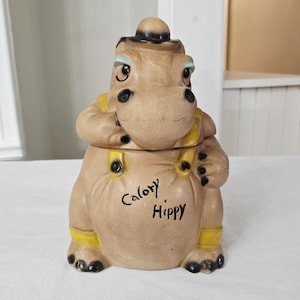 May include: Tan ceramic cookie jar shaped like a cartoon hippo wearing a hat and overalls. The jar has black accents and the words "Calory Hippy" are written on the front.