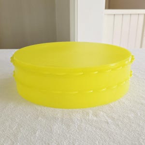 May include: A bright yellow, round container with a lid. The container has a scalloped edge design and is made of a solid, opaque material. The container is sitting on a white surface.