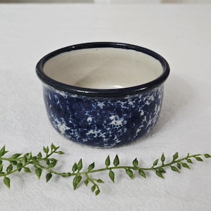 May include: A small, round ceramic bowl with a speckled navy blue exterior and a cream-colored interior. The rim is a solid navy blue. The bowl is sitting on a white surface with a green leafy vine in the foreground.