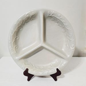 May include: A cream-colored, divided serving dish with three sections. The round plate features a textured surface and is decorated with a raised, floral vine pattern around the rim. The dish is displayed on a dark brown stand.