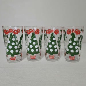 May include: Four clear drinking glasses with Christmas tree designs. Each glass features a green Christmas tree with white dots, red poinsettias, and holly leaves. The glasses have a festive, vintage aesthetic.
