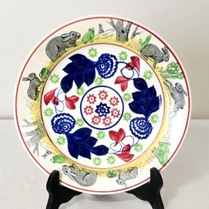 May include: A decorative ceramic plate featuring a whimsical design. The plate's border showcases rabbits and frogs in a pastoral scene. The center has blue and red floral patterns. The plate is supported by a black stand.