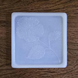 May include: Square, light blue coaster with a raised floral design. The coaster features a detailed embossed pattern of a flower and leaves. The edges are slightly raised, providing a defined border. The coaster is made of a solid, opaque material.