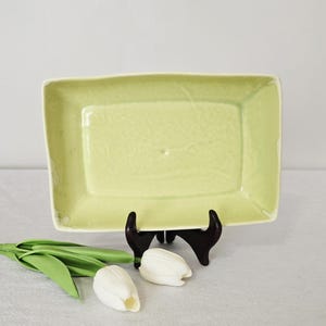 May include: A rectangular, pale green ceramic serving dish with a slightly irregular edge. The dish is displayed on a dark brown wooden stand. White tulip buds and green leaves are arranged nearby, suggesting a spring theme.