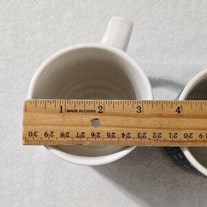 May include: Two white ceramic mugs viewed from above, with a wooden ruler placed across the top of the left mug. The ruler shows the mug opening has a diameter of approximately 3.5 inches. The mugs have a smooth, plain interior and a handle.