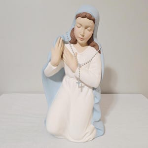 May include: A kneeling statue of a woman in a white dress and a light blue veil. The statue has a rosary and is in a prayerful pose with closed eyes. The statue is on a white surface.