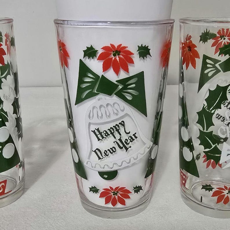 May include: Set of three clear drinking glasses with a festive New Year's design. Each glass features a white background with green bow, red poinsettias, and the text "Happy New Year" in black script. The glasses are ideal for holiday celebrations.