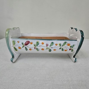 May include: A white ceramic planter in the shape of a cradle, featuring a hand-painted floral design in green, orange, and red. The planter has a rectangular interior and curved ends with green accents. The cradle planter is a decorative item.