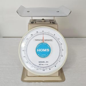 May include: Vintage beige and white kitchen scale with a stainless steel tray. The scale face features a blue and white logo and reads "HOMS MODEL 20 20 lbs x 1 oz". The dial has a red needle and temperature compensation text.