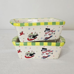 May include: Two rectangular ceramic baking dishes. The dishes are off-white with a yellow and green striped rim. The sides are decorated with images of a penguin on a sled, a snowman on skis, red birds, and snowflakes.