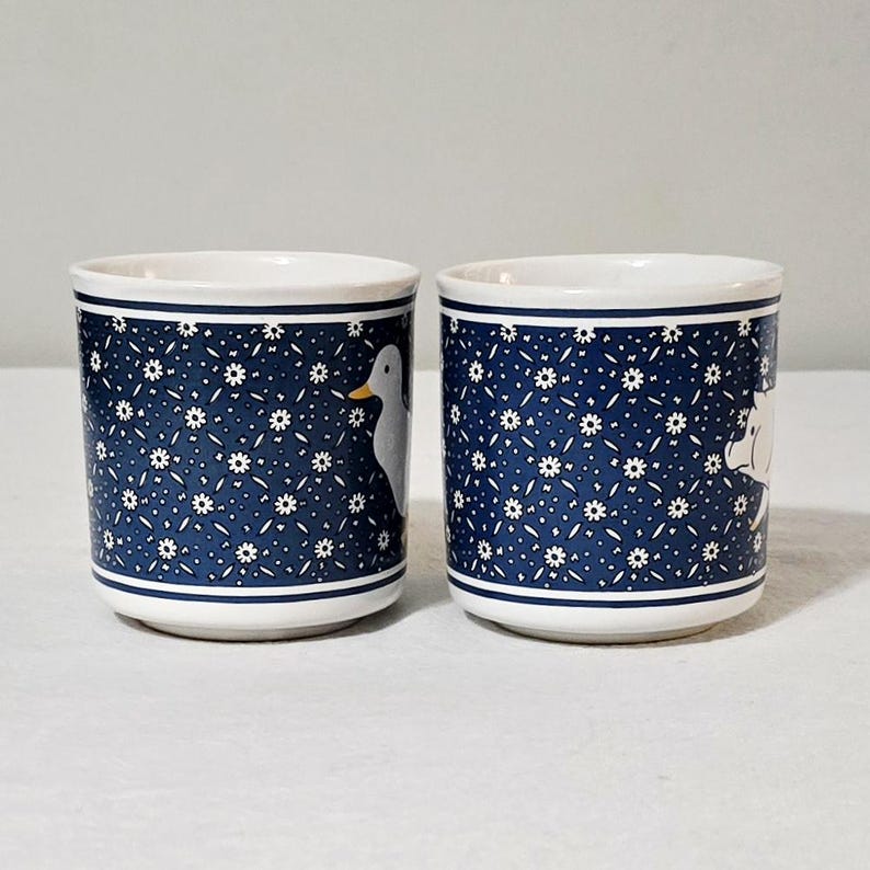 May include: Two cylindrical ceramic mugs featuring a deep blue background with a repeating white floral pattern. One mug displays a white duck with a yellow beak, while the other mug features a white pig. Both mugs have white rims and bases.