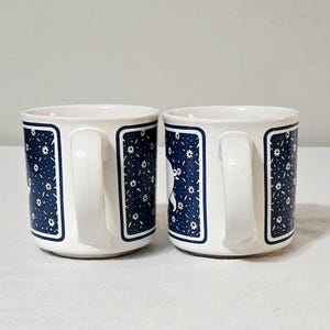 May include: Two white ceramic mugs featuring a dark blue rectangular panel on the side. The blue panels are decorated with a repeating pattern of small white flowers and thin white lines. Each mug has a curved handle and a smooth, rounded base.