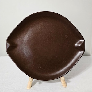 May include: A brown, square-shaped ceramic plate with rounded corners. The plate has a slightly textured surface and is displayed on a small wooden stand. The plate's color is a deep, rich brown.
