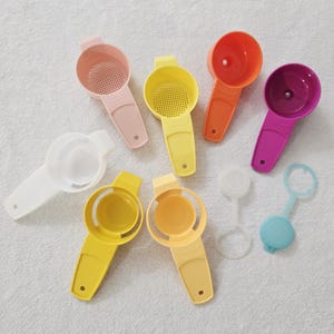 May include: Assortment of colorful kitchen tools, including egg separators, strainers, and measuring spoons. The tools are in shades of pink, yellow, orange, purple, white, and blue. The tools are made of plastic and are arranged on a white surface.