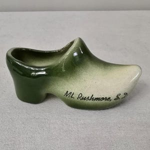 May include: Green and white ceramic clog with the words "Mt. Rushmore, S.D." written on the side.