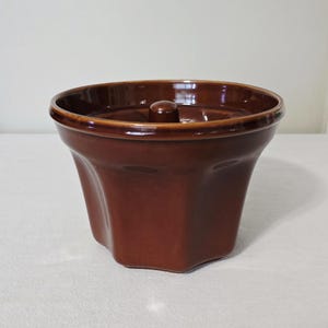 May include: A brown ceramic bundt pan with a rounded rim and a central chimney. The pan has a square base that curves inward and then outward to form a wider top. The pan is a solid, glossy brown color.
