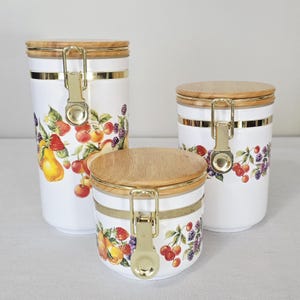 May include: Set of three white ceramic canisters with wooden lids and gold-tone accents. Each canister features a colorful fruit design with pears, strawberries, cherries, and blackberries. The canisters have a metal clasp closure.