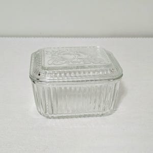 May include: A clear rectangular glass butter dish with a lid. The base features vertical ribbed detailing along the sides. The lid has a matching ribbed border and a central embossed design depicting a cluster of grapes and leaves.
