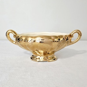 May include: A gold-colored, decorative bowl with two handles. The bowl has a scalloped rim and sits on a small, ornate base. The interior of the bowl is a cream color, contrasting with the shiny gold exterior.