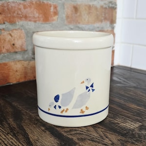 May include: A cream-colored ceramic utensil holder with a blue and tan goose design. The cylindrical holder has a blue stripe near the base and is set on a dark wooden surface. The geese are wearing blue bow ties.