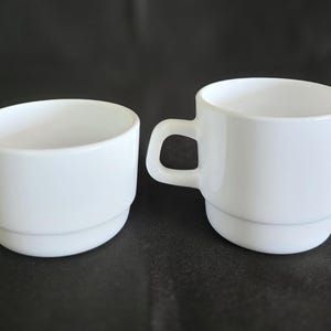 May include: Two white ceramic cups, one with a handle and the other without. Both cups have a simple, rounded design.