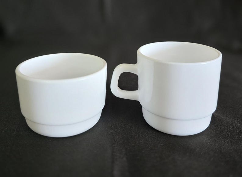 May include: Two white ceramic cups, one with a handle and the other without. Both cups have a simple, rounded design.