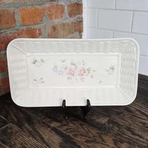 May include: An off-white ceramic serving platter with a woven basket design around the edges. The centre has a floral pattern with pink, blue, and green flowers. The platter is displayed on a black stand.