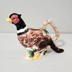 May include: A ceramic pheasant-shaped creamer with a black head, red facial markings, and a yellow beak. The body is brown with feather detailing, and the handle is designed to look like a tree branch. Green leaves and yellow feet add to the decorative design.
