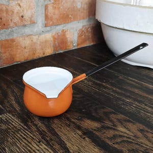 May include: An orange enamel saucepan with a long black handle. The interior is white, and the handle is attached to the side of the pan. The saucepan is on a dark wooden surface, with a brick wall in the background.