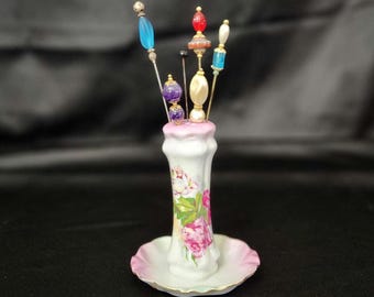 May include: A white porcelain pin cushion holder with a pink floral pattern and a scalloped base. Five decorative hat pins with colourful glass and pearl-like beads in shades of blue, purple, red, and cream are inserted into the top opening.