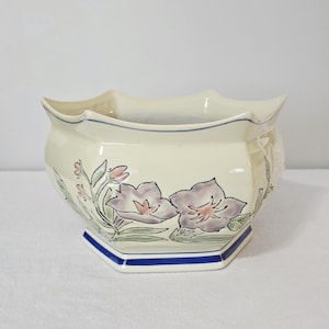 May include: Cream ceramic planter with a scalloped rim and a hexagonal base. It features hand-painted lavender flowers with green leaves and a blue stripe. A decorative piece for the home, suitable for plants.