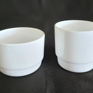 May include: Two white ceramic cups, one taller than the other, both with a simple, rounded design.