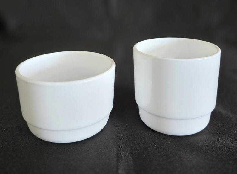 May include: Two white ceramic cups, one taller than the other, both with a simple, rounded design.