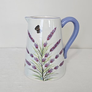 May include: White ceramic pitcher with a lavender and butterfly design. The pitcher features a light blue handle and is decorated with purple lavender sprigs and a black butterfly. A decorative kitchenware item.