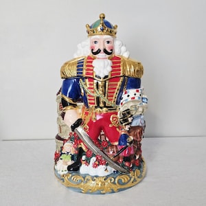 May include: A ceramic nutcracker cookie jar featuring a central figure in a red and blue uniform with a gold crown and sword. The base is decorated with small figures, including a child, a soldier on a horse, and white swans amidst red flowers.