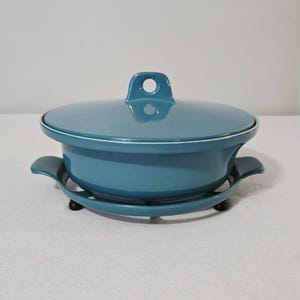 May include: A teal-colored, lidded casserole dish with a matching trivet. The dish has a rounded shape with a white rim and a handle on the lid. The trivet has curved handles and small black feet. A kitchenware item.